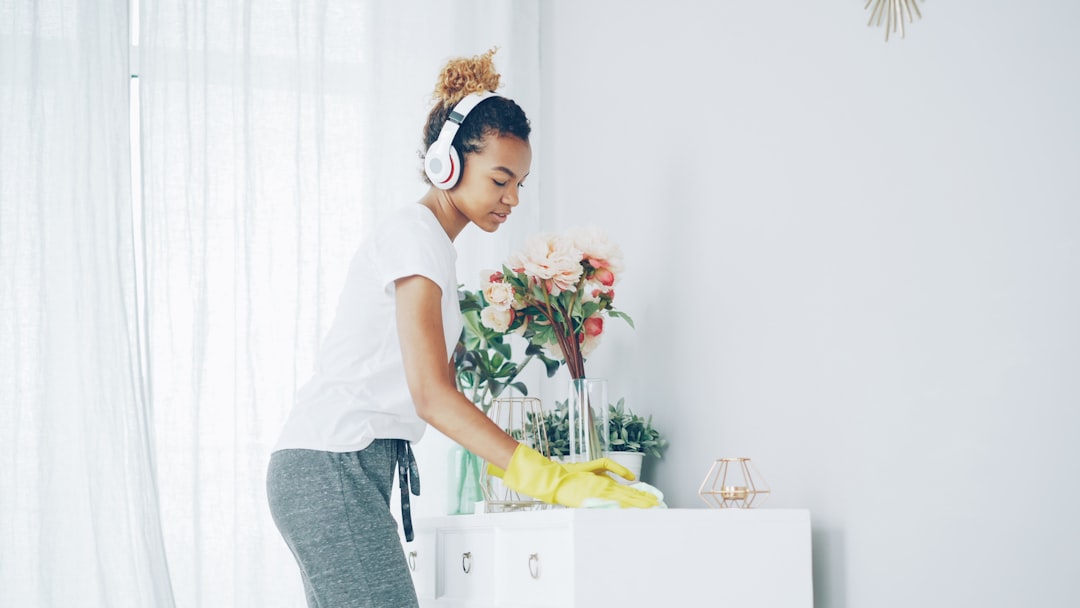 Cheerful maid African American girl is doing housework dusting using cloth and wearing protective gloves, young woman is listening to music in headphones